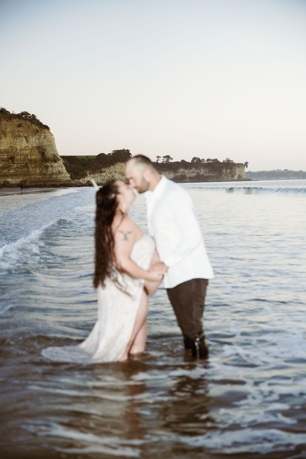 A couple embraces in shallow ocean waters during a romantic beach photo session at sunset.