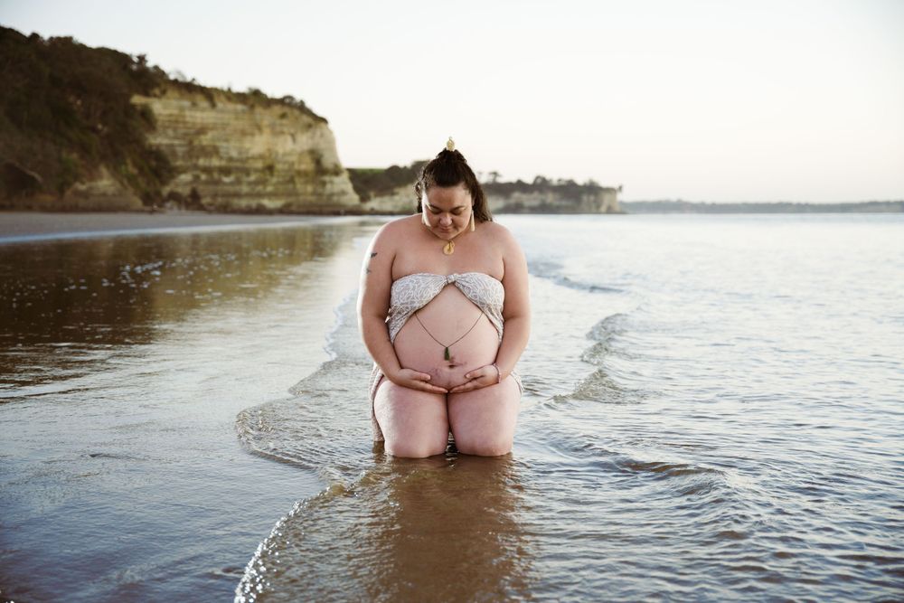 Person wearing white sits in shallow ocean waters during a beach maternity photography session.