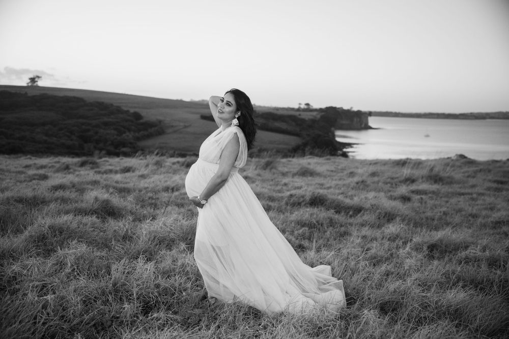 Pregnant woman in flowing white dress poses against coastal landscape.