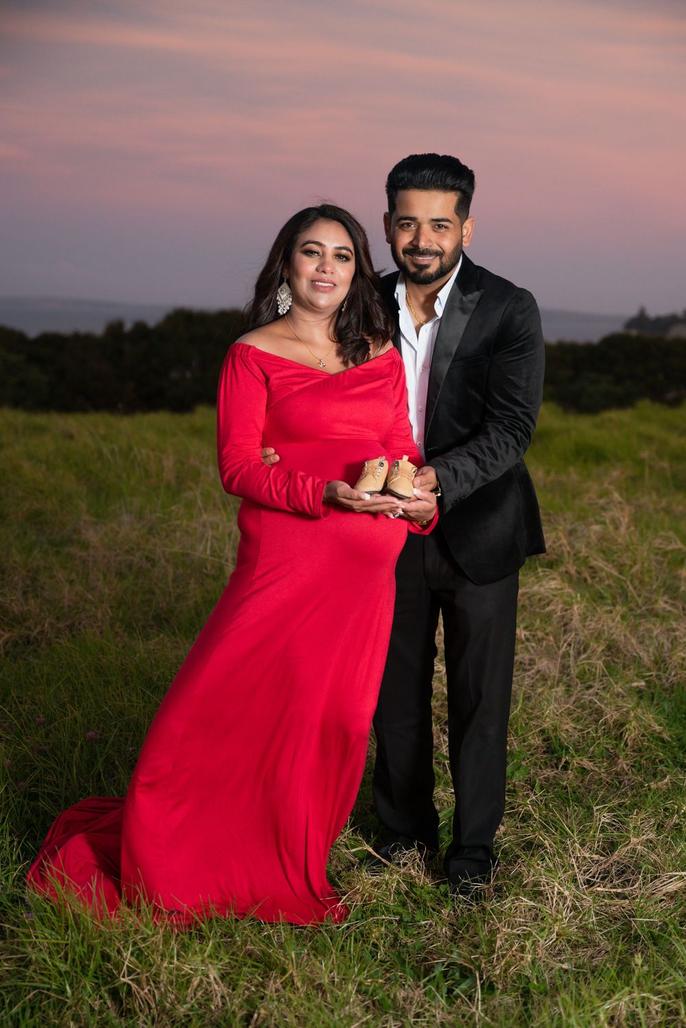 Couple in formal attire posing in field during sunset with red dress flowing.
