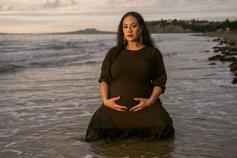 Person in black dress poses for maternity photos while sitting in shallow ocean waves at sunset.