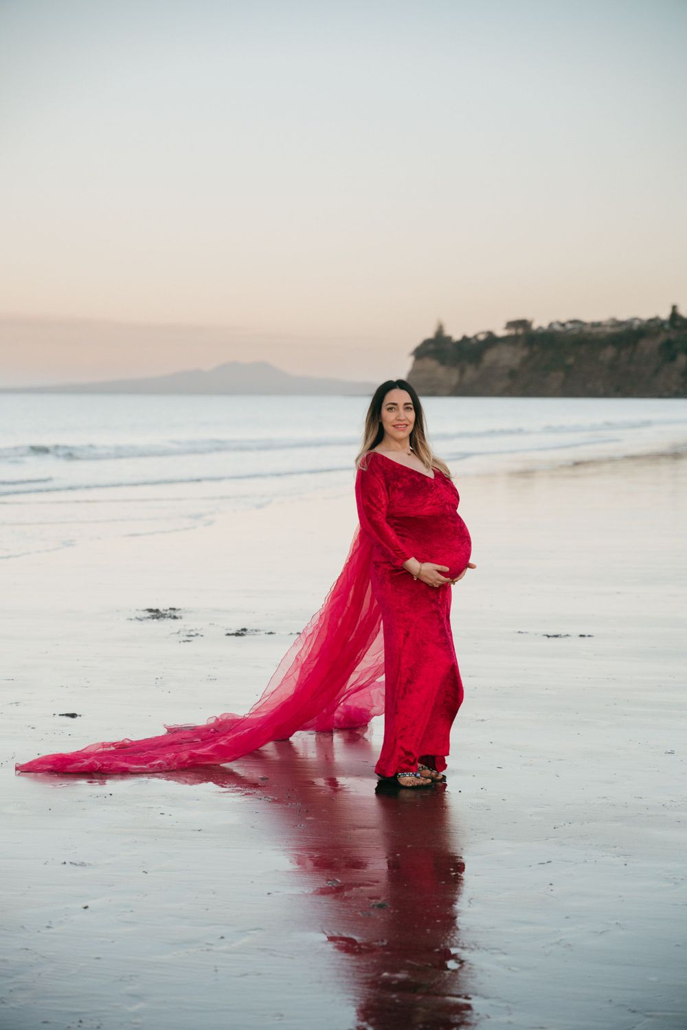 A flowing red dress catches the wind during a sunset maternity photoshoot on a beach with coastal cliffs.