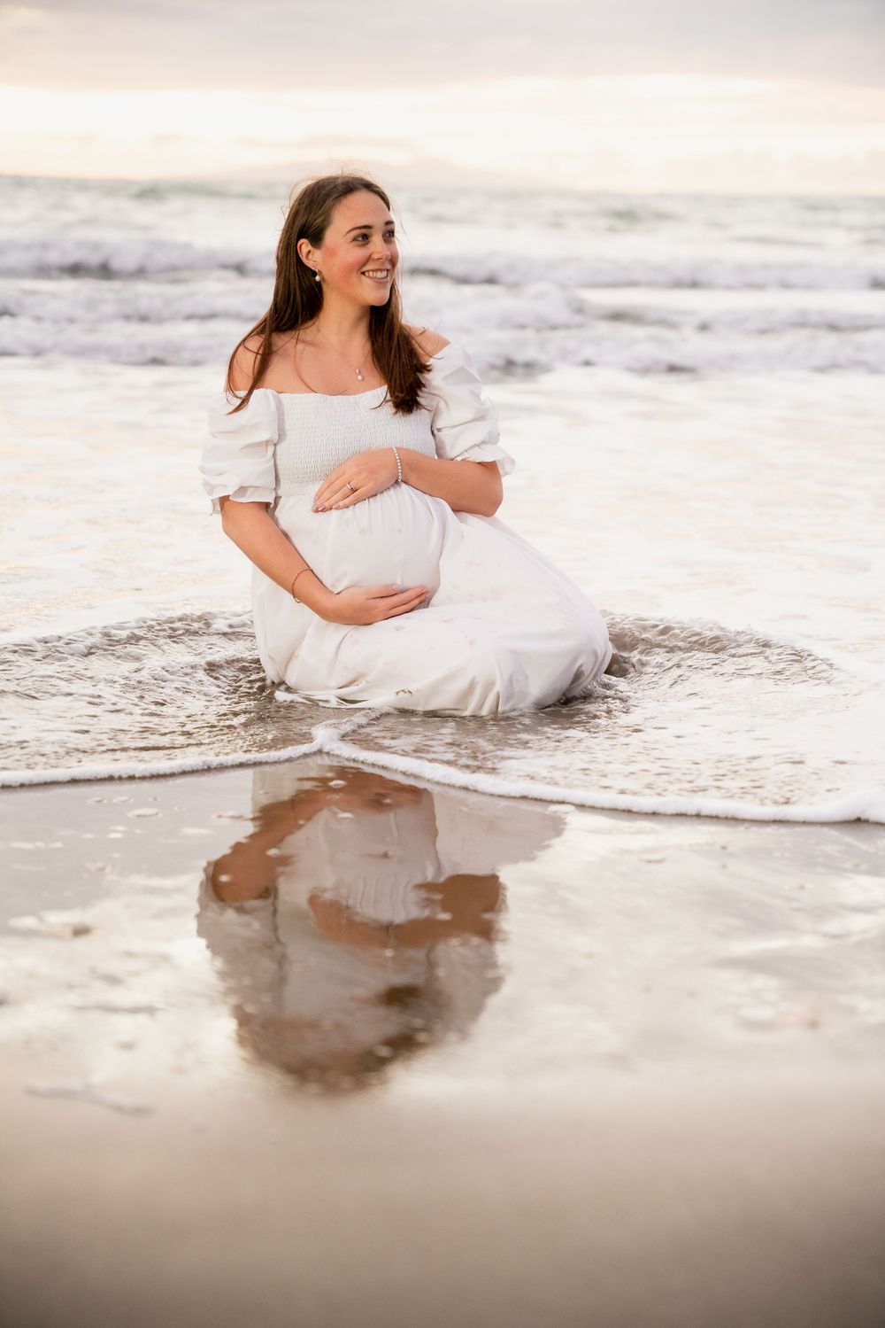 Person in white off-shoulder dress sits in ocean waves during sunset beach maternity shoot.