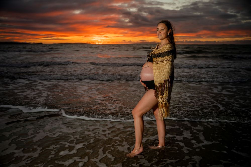 Person in gold wrap poses on beach during dramatic sunset maternity photoshoot.