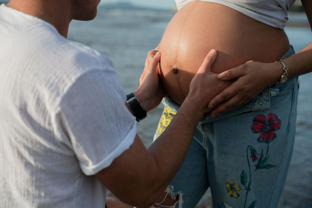 Close-up of hands cradling pregnant belly against ocean backdrop.