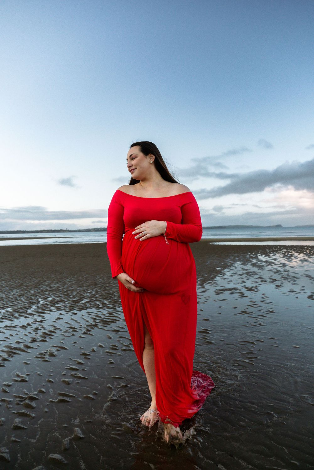 Person in flowing red dress stands in shallow waters during beach maternity photoshoot.