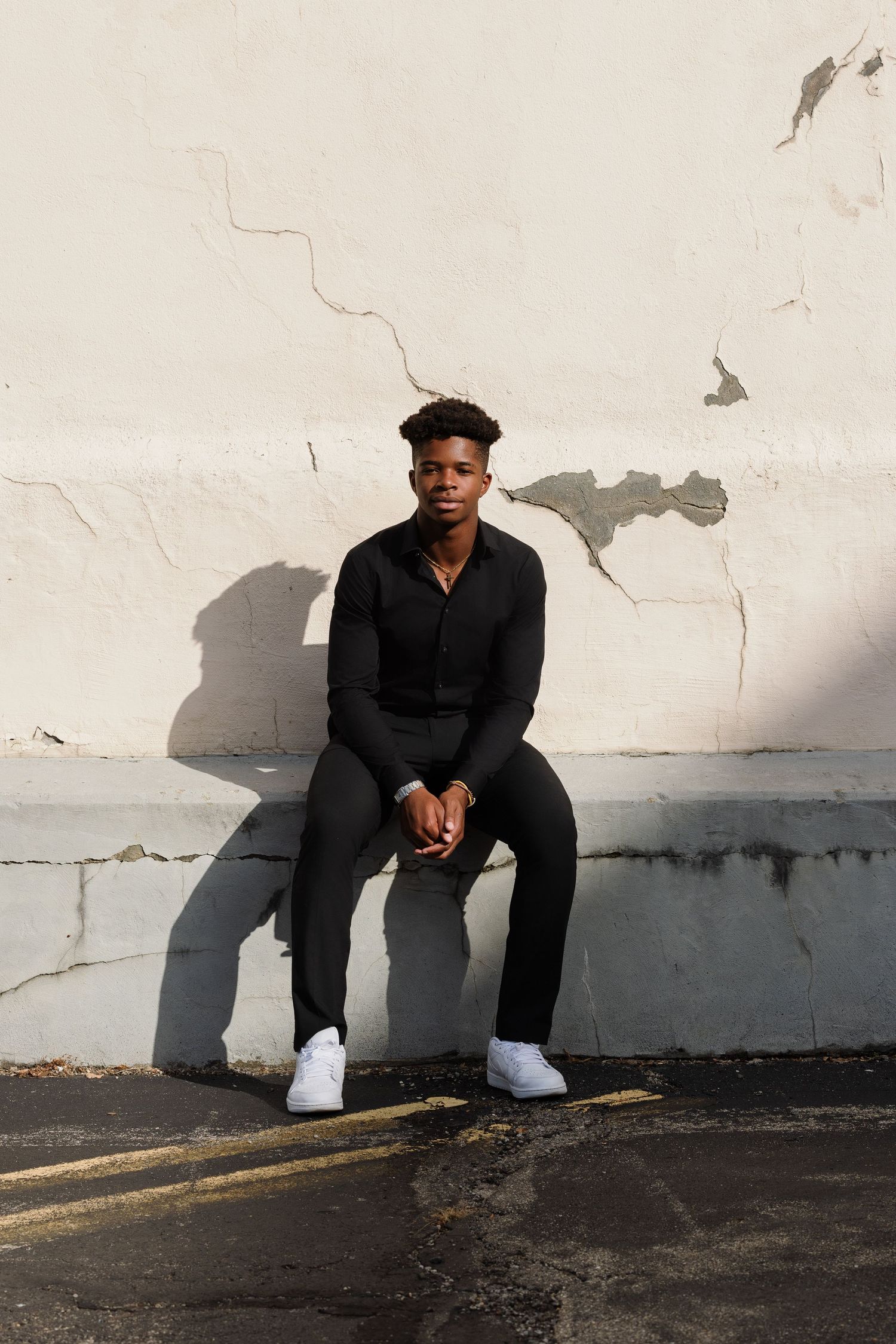 A person in black attire sits against a white wall creating a striking shadow contrast.