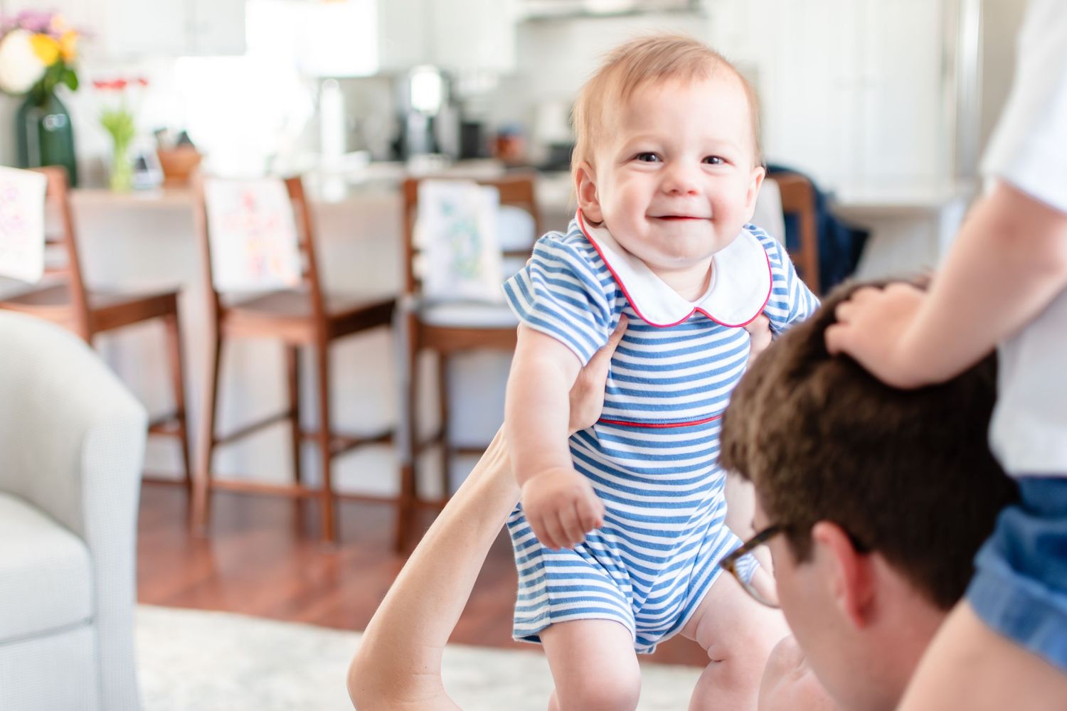 A joyful baby in striped outfit being lifted up in a living room setting.