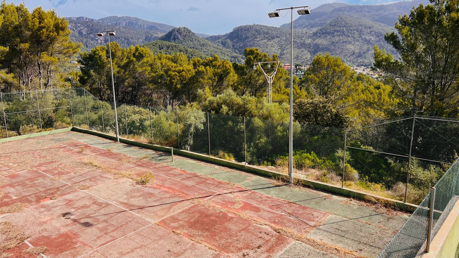 Tennis court surrounded by trees with mountain views in Mediterranean countryside setting.