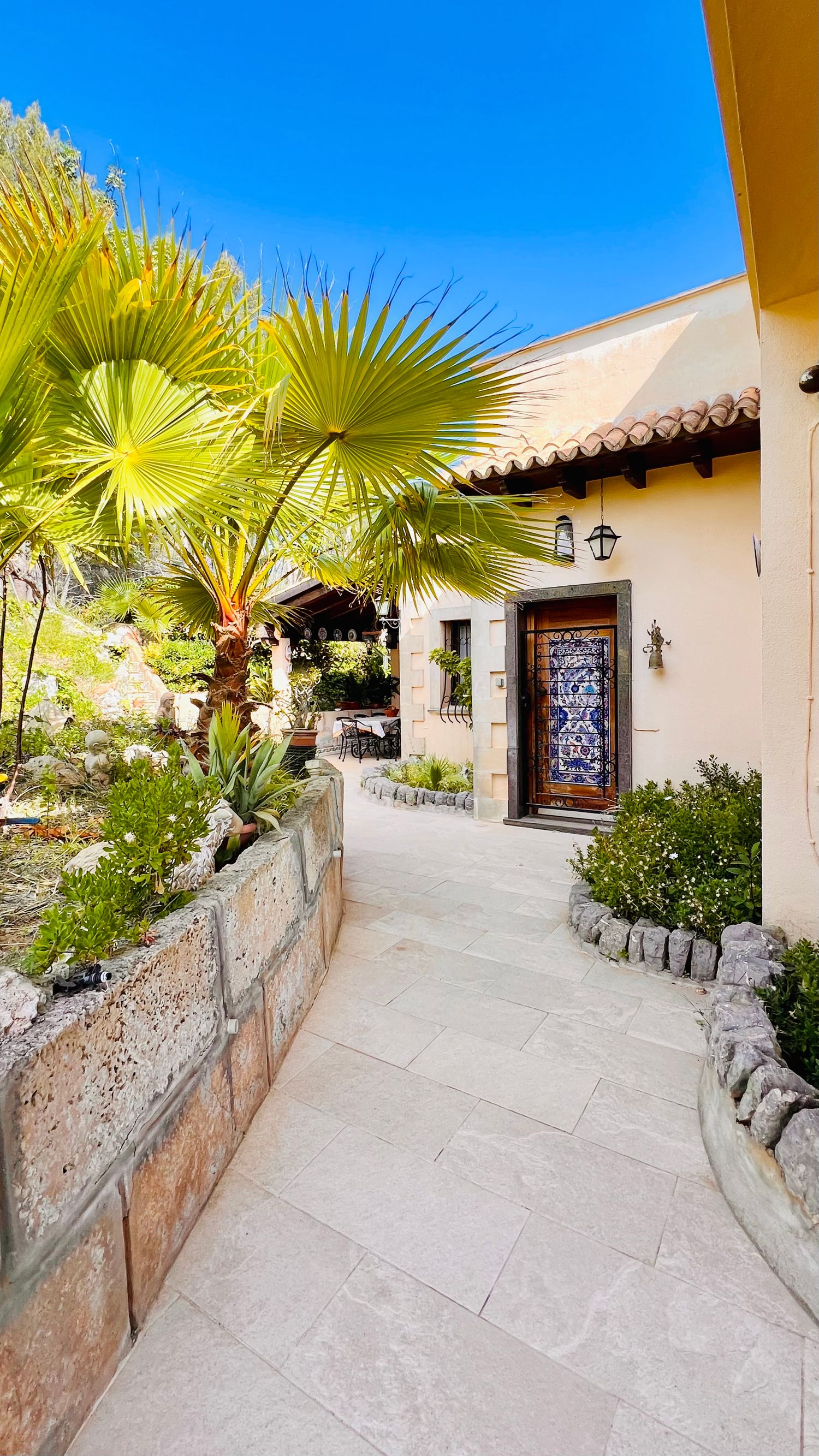 Curved stone pathway leading to Spanish-style entrance with palm trees and decorative door.