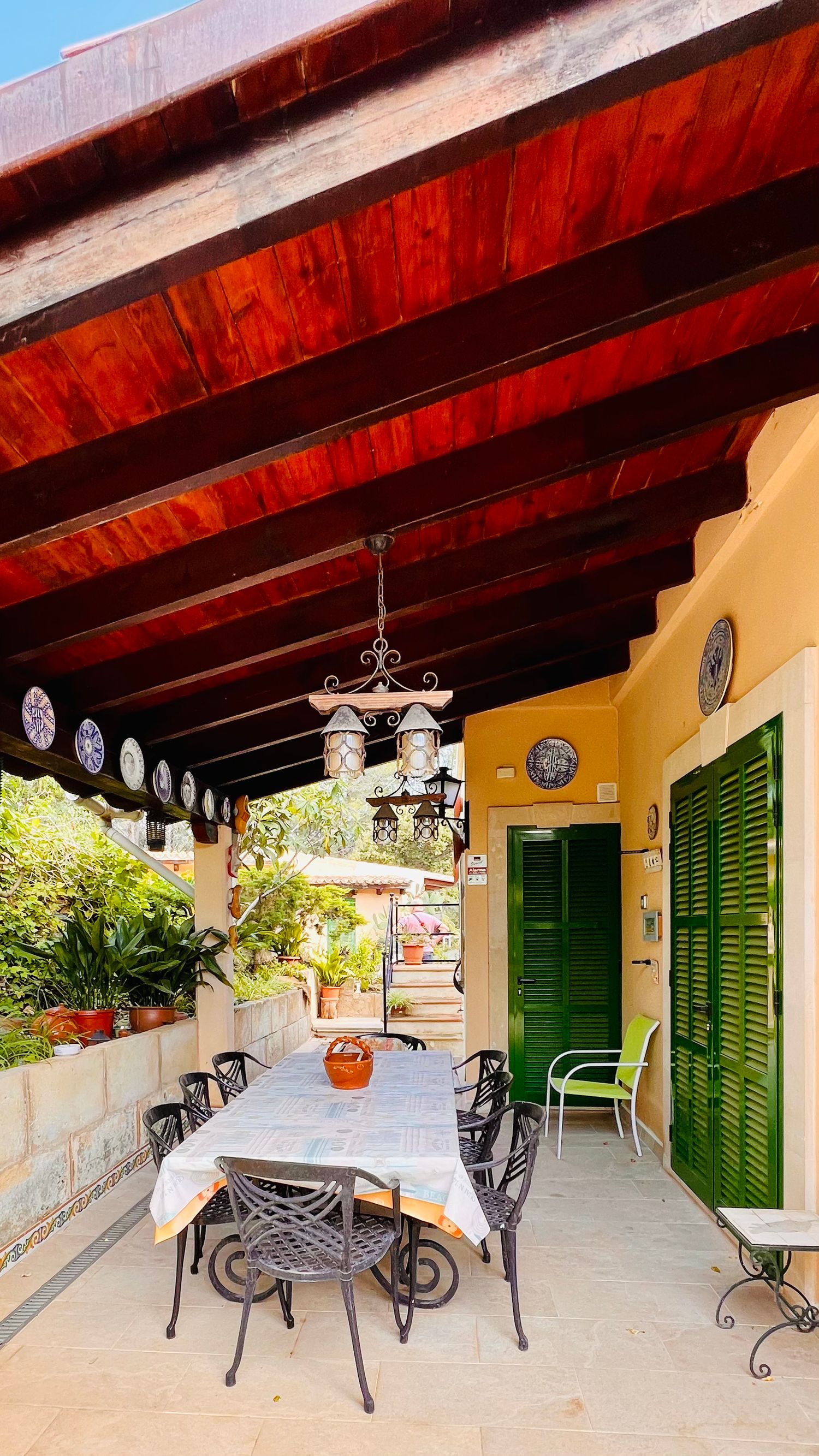 Covered patio dining area with wooden beams, green shutters, and decorative hanging lamps.