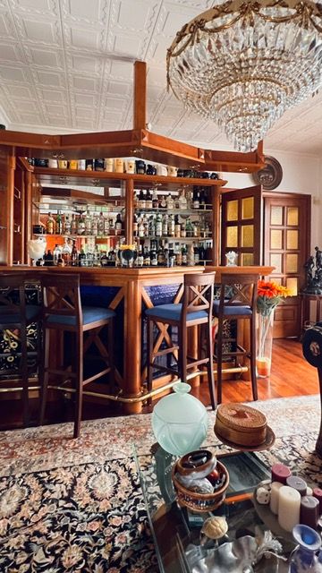 Traditional home bar with crystal chandelier, wooden cabinetry, and bar stools beneath ornate ceiling.