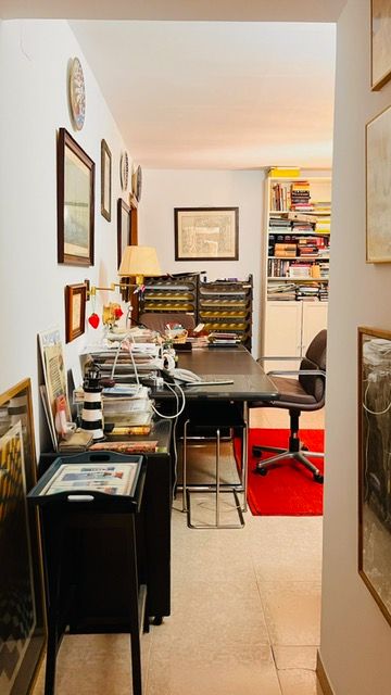 Home office space with desk setup, filing cabinets, and artwork-lined walls leading to book-filled shelves.