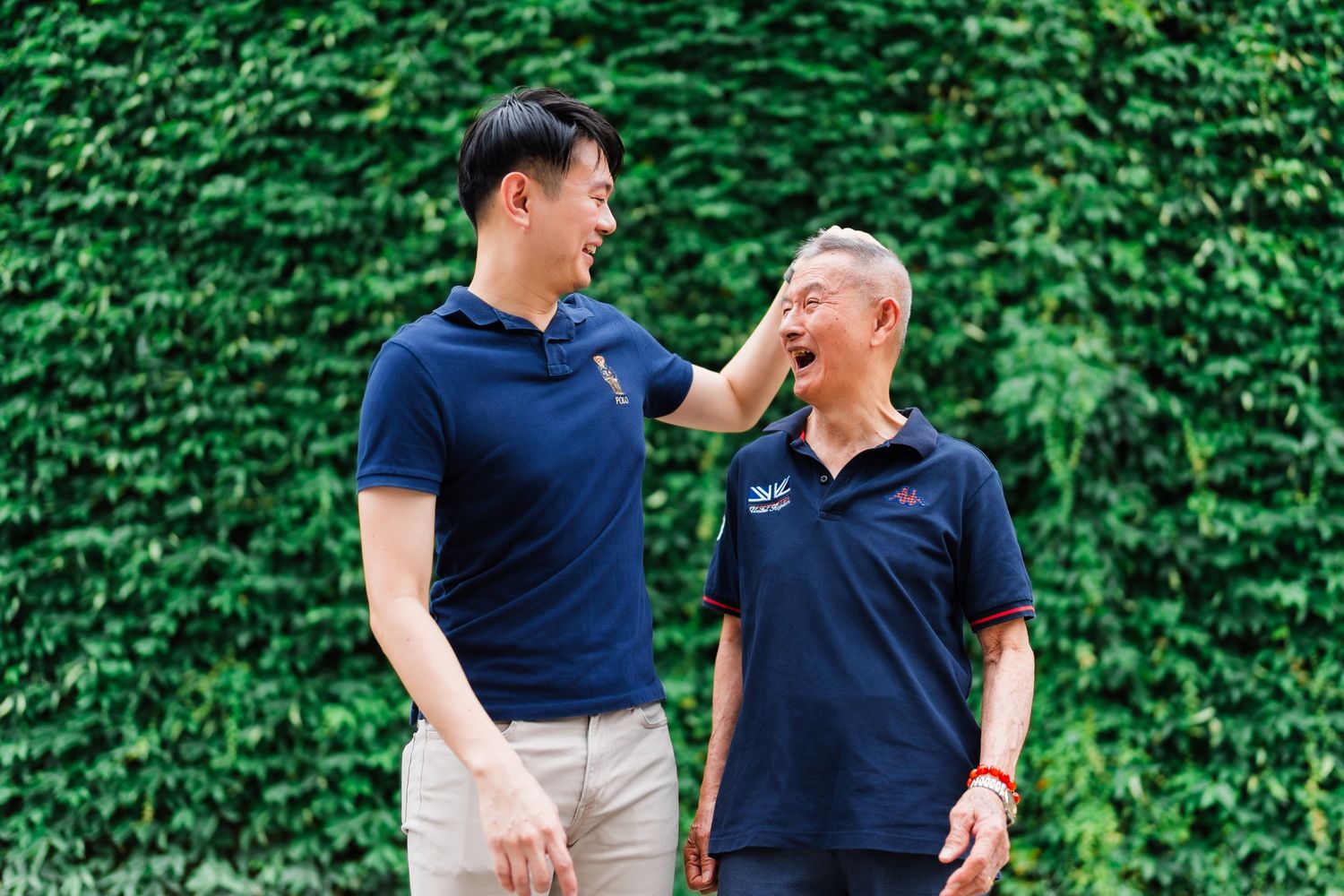 Two men in navy blue polo shirts sharing a laugh against ivy wall background.