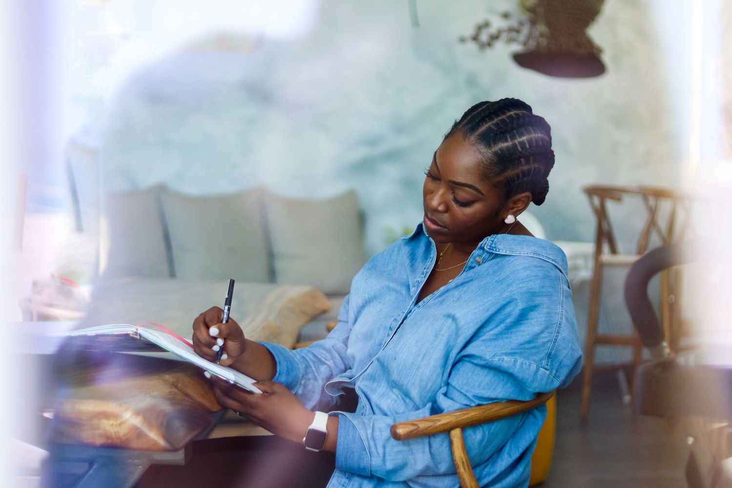 Person in blue shirt writing in notebook while studying at desk.