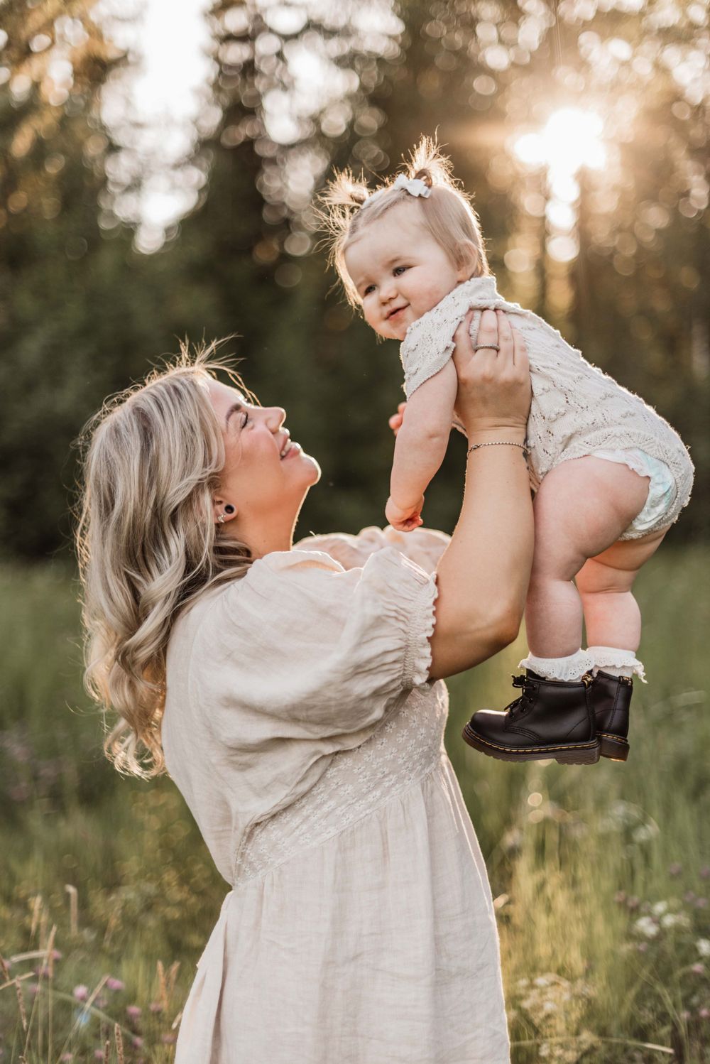 Woman in white dress holds up baby in lace outfit against golden sunset in outdoor nature setting.