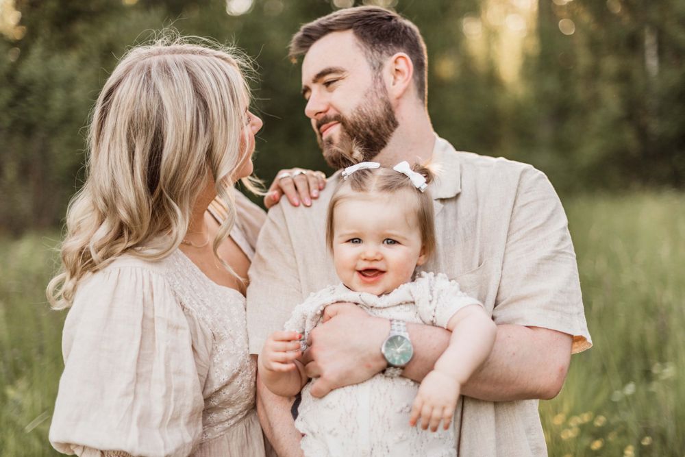 Three people in soft neutral colored clothing share a joyful moment together outdoors in a sunlit meadow.