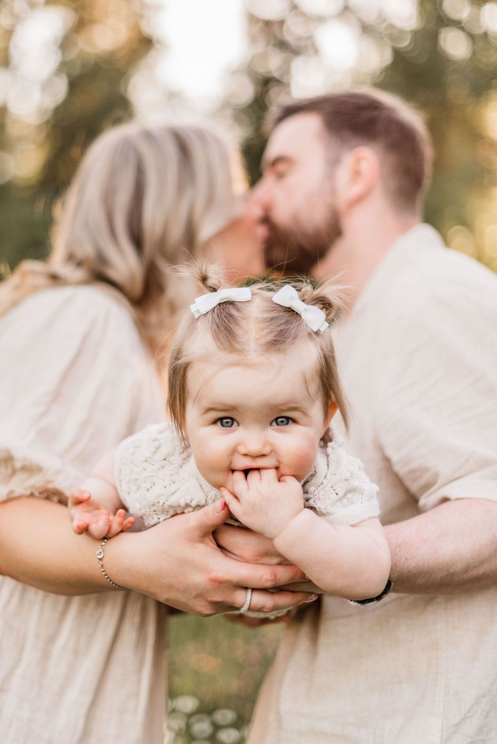 A sweet baby in white dress smiles while held by family during a warm outdoor photo session.