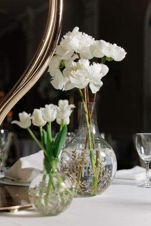 White peonies and tulips in glass vases as table centerpieces at an elegant dinner.