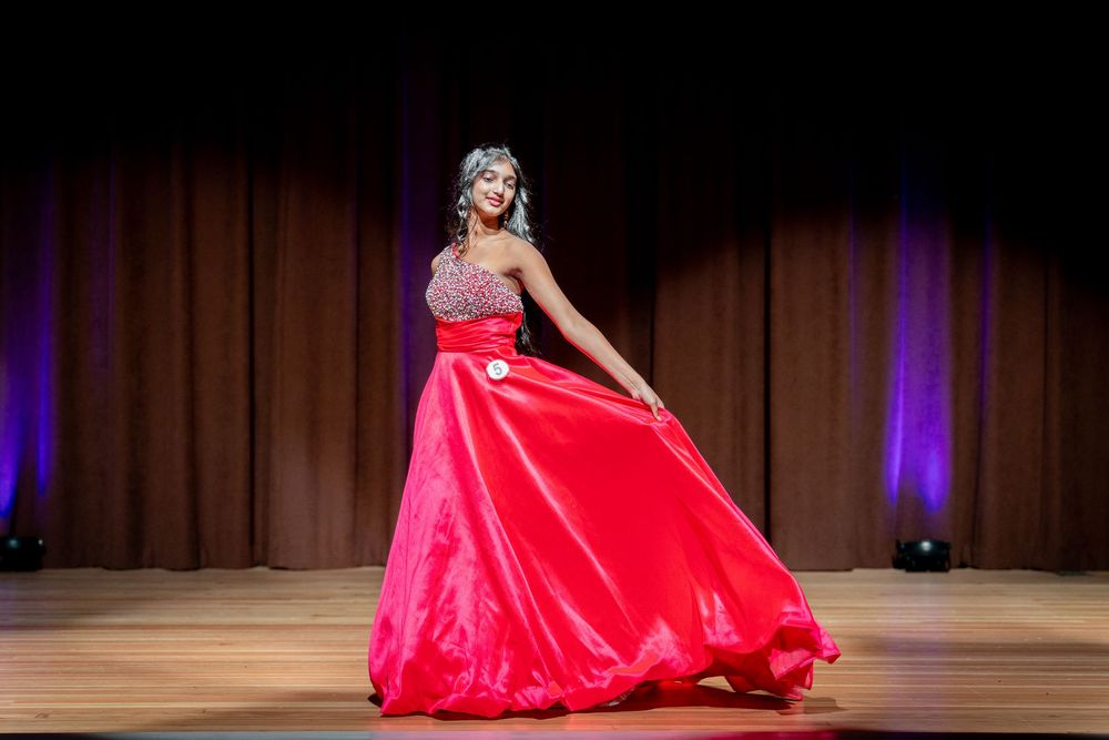 Woman in red ballgown twirling on stage during a pageant competition under purple stage lights.