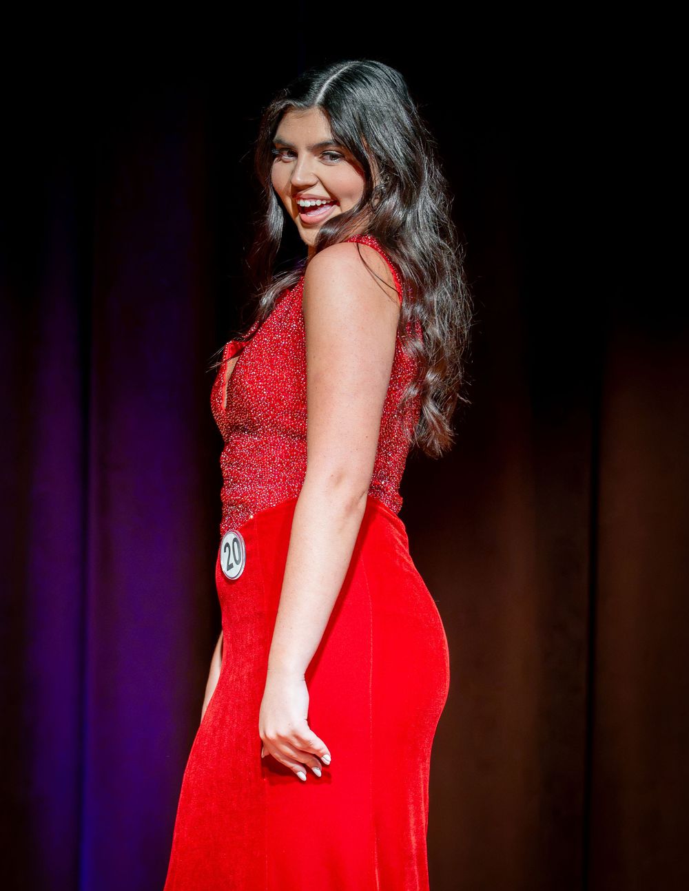 Woman in red velvet gown smiling over shoulder on stage at beauty pageant competition.