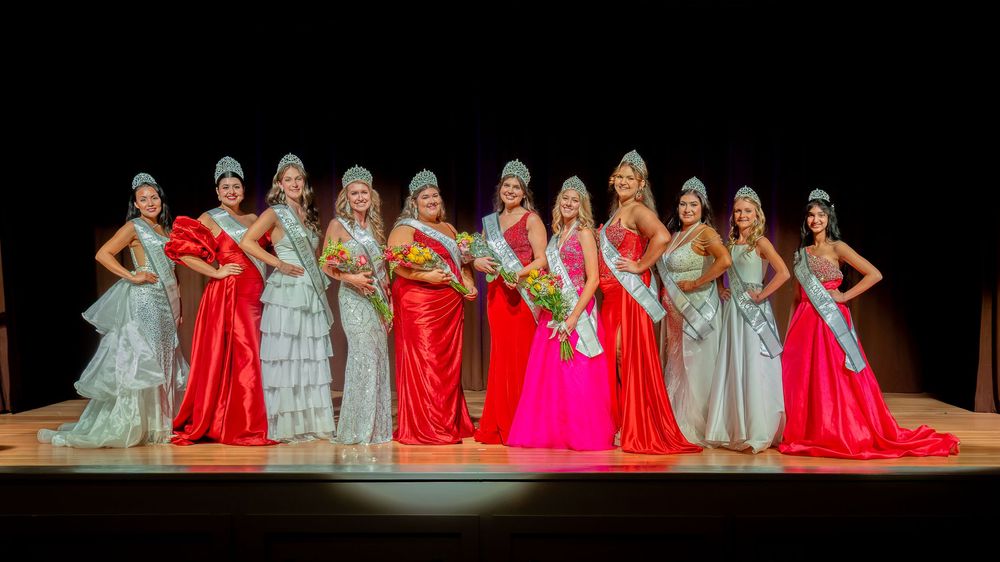 A group of beauty pageant contestants in elegant gowns and tiaras pose together on stage under dramatic lighting.