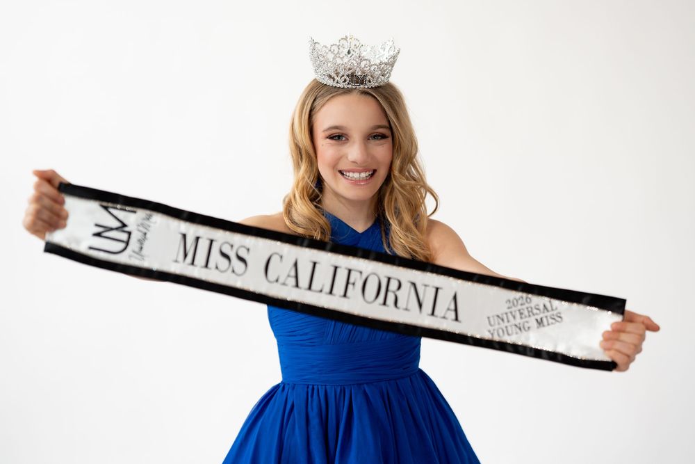 A pageant contestant in a blue evening gown holds up a sash while posing for a photo shoot.