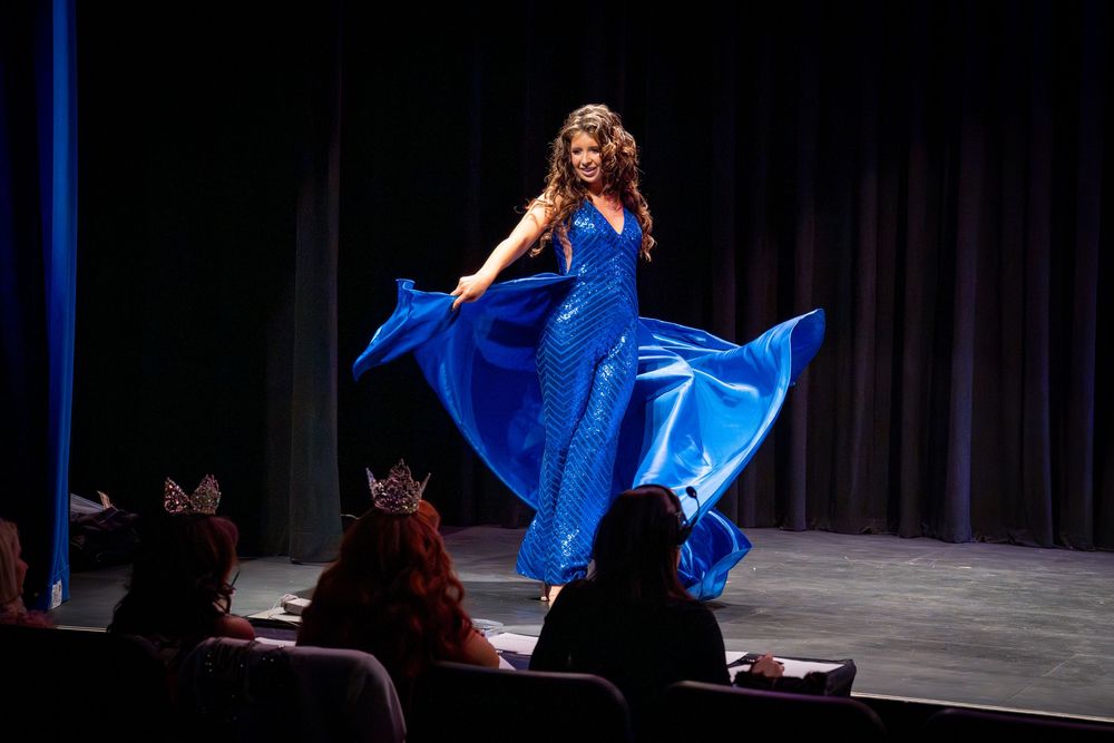 A performer in a flowing royal blue gown twirls gracefully on stage while an audience watches in silhouette.