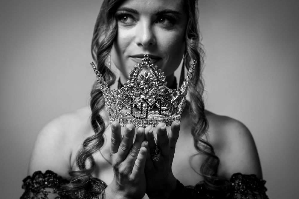 Black and white portrait of a woman holding an ornate jeweled crown with both hands, gazing upward.
