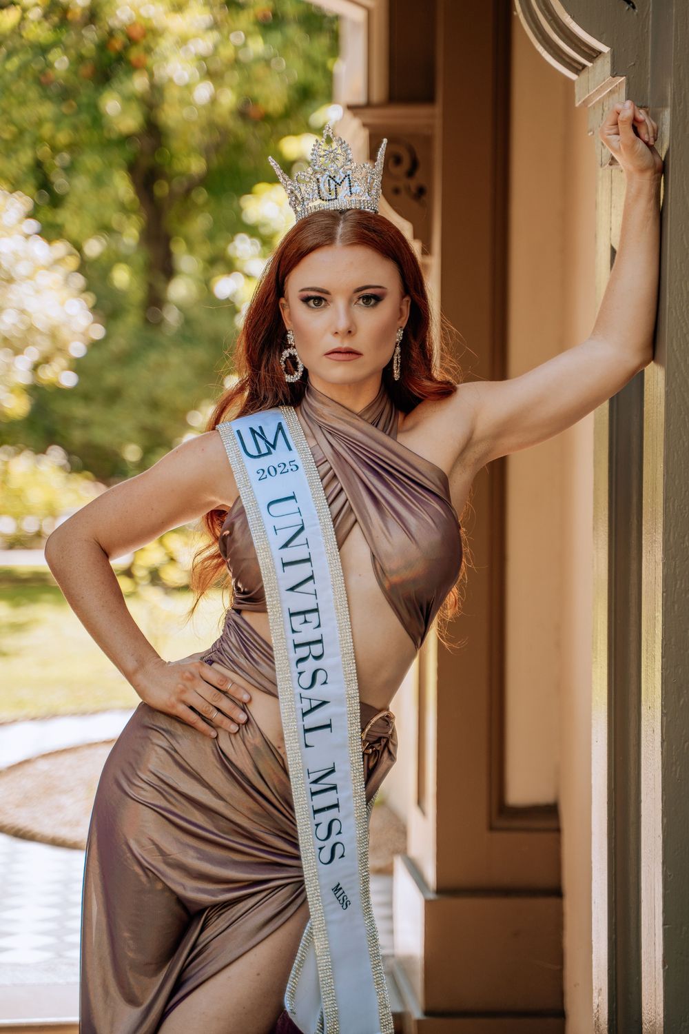 A pageant contestant wearing a shimmering bronze gown and crown poses dramatically against a pillar in warm sunlight.