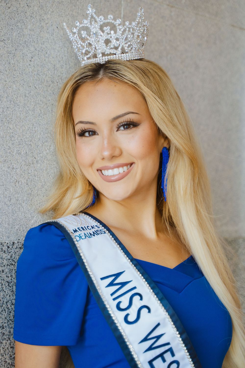 Pageant contestant wearing a royal blue dress and sparkling crown poses against grey backdrop with bright smile.
