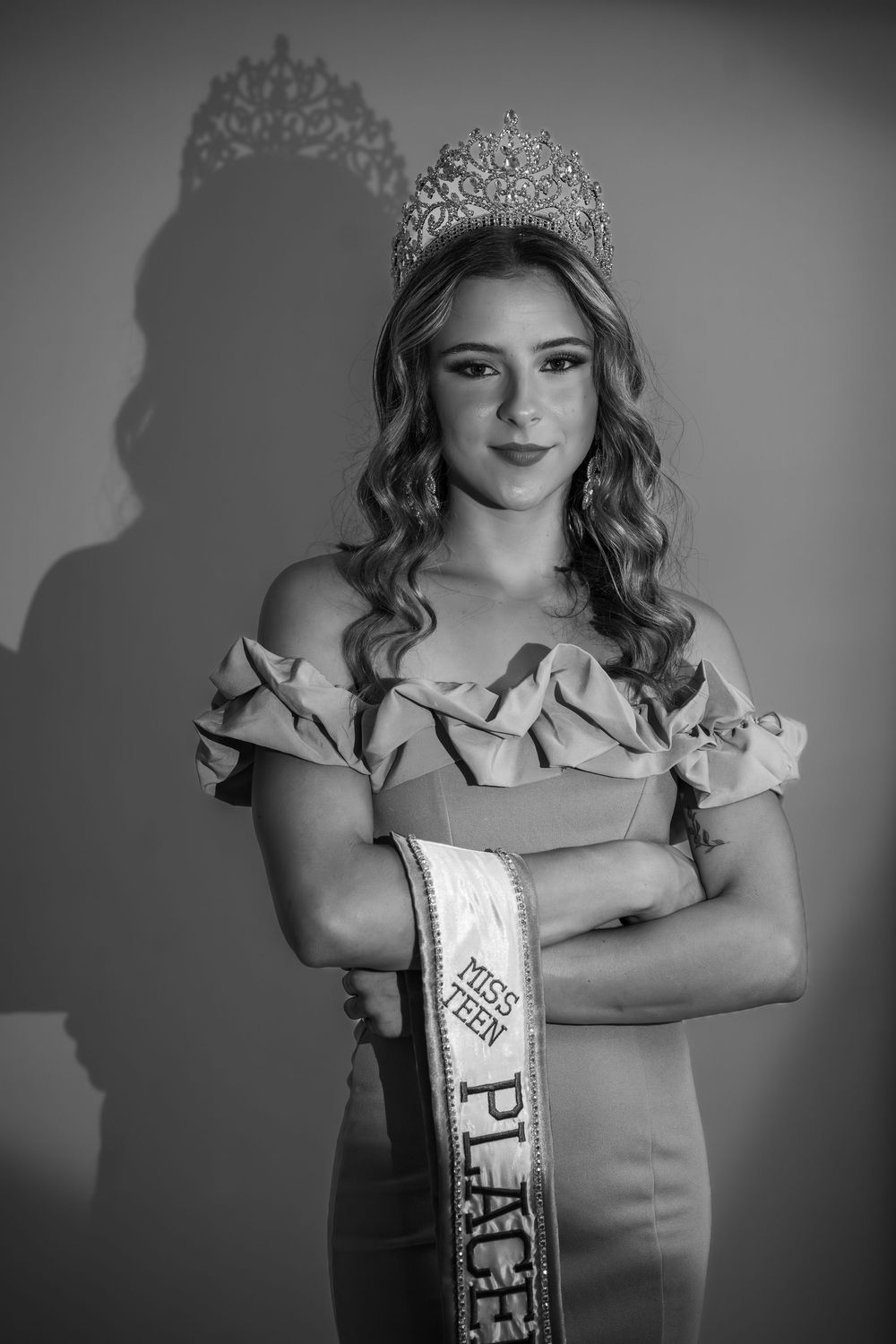 Black and white portrait of a pageant contestant wearing an ornate tiara, sash and ruffled formal gown.