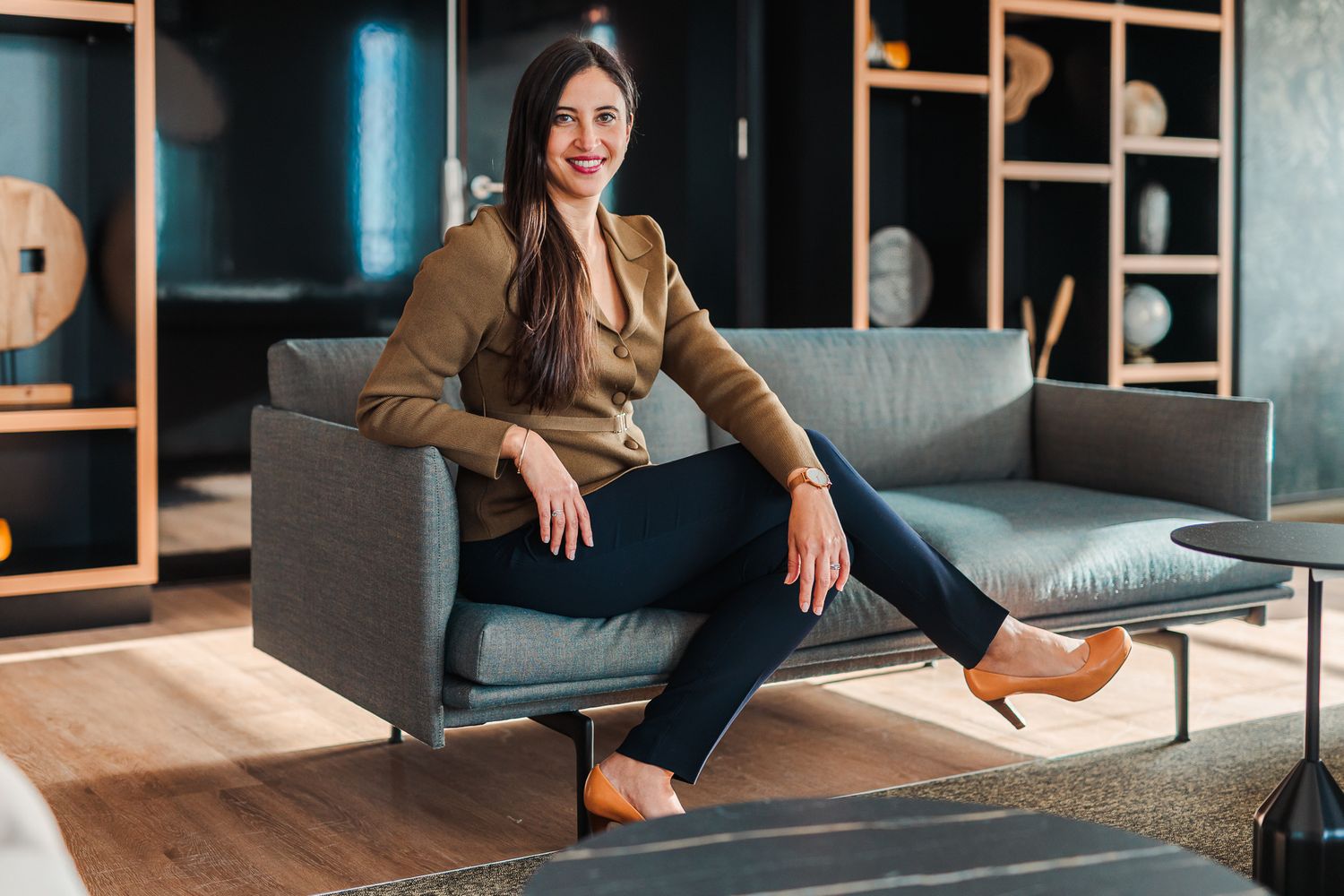 Young brunette woman in blue shirt and white pants sits thoughtfully on a sofa in a modern home.