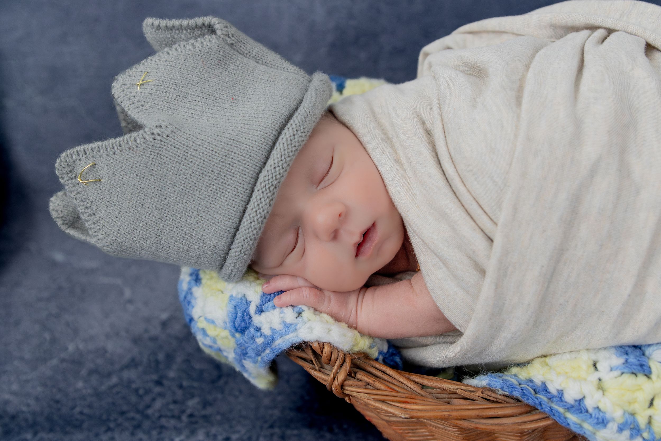 Sleeping newborn wearing a gray knit hat rests in a cozy basket with blue and yellow accents.