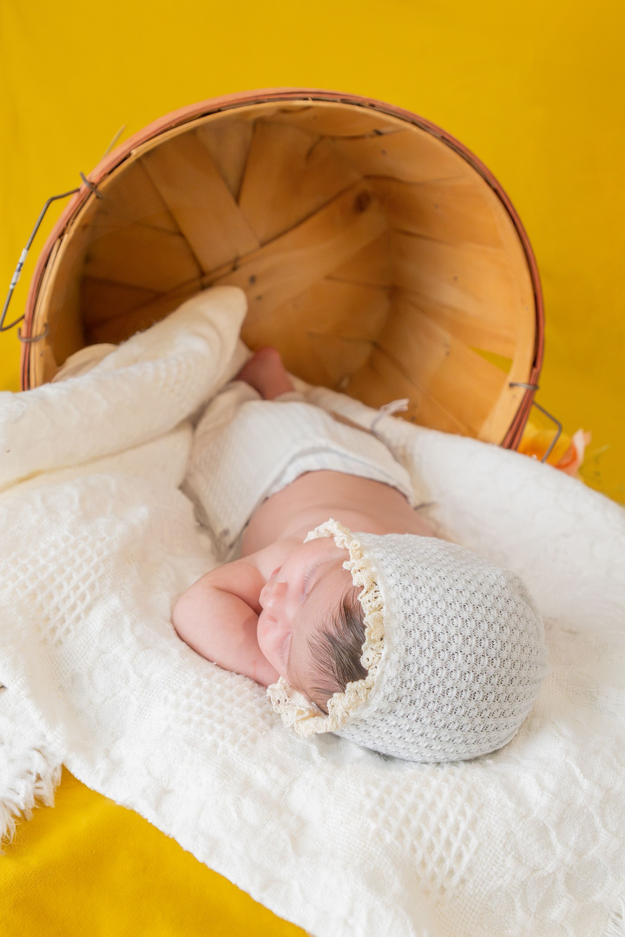 Newborn in knit bonnet sleeping peacefully in wooden basket against yellow background.
