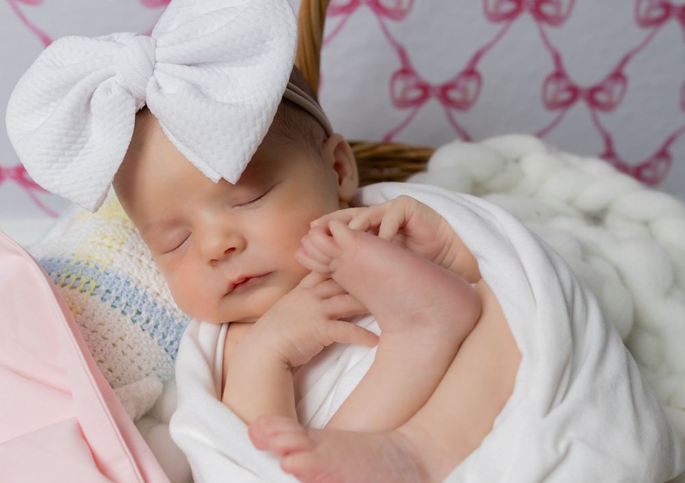 Sleeping newborn baby girl wearing a white bow headband, swaddled in white, with tiny hands visible.
