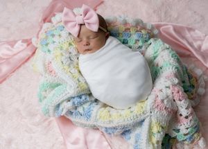 Newborn baby girl swaddled in white wrap, wearing pink bow headband, resting on colorful pastel crochet blanket.