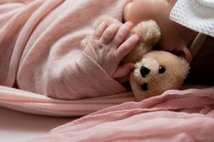 Baby hand holding a small teddy bear under a soft pink blanket, cozy and peaceful nursery scene.