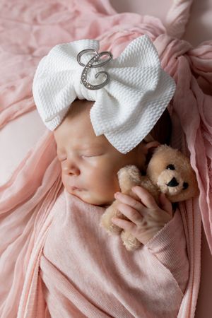 Sleeping newborn baby girl wrapped in pink swaddle, wearing white bow headband with C initial, holding tiny teddy bear.