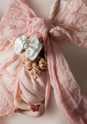 Newborn baby swaddled in pink wrap with bow, holding tiny teddy bear, wearing white monogram bow headband.