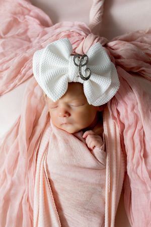 Newborn baby girl swaddled in pink wrap, sleeping peacefully wearing a white bow headband with crystal letter C initial.