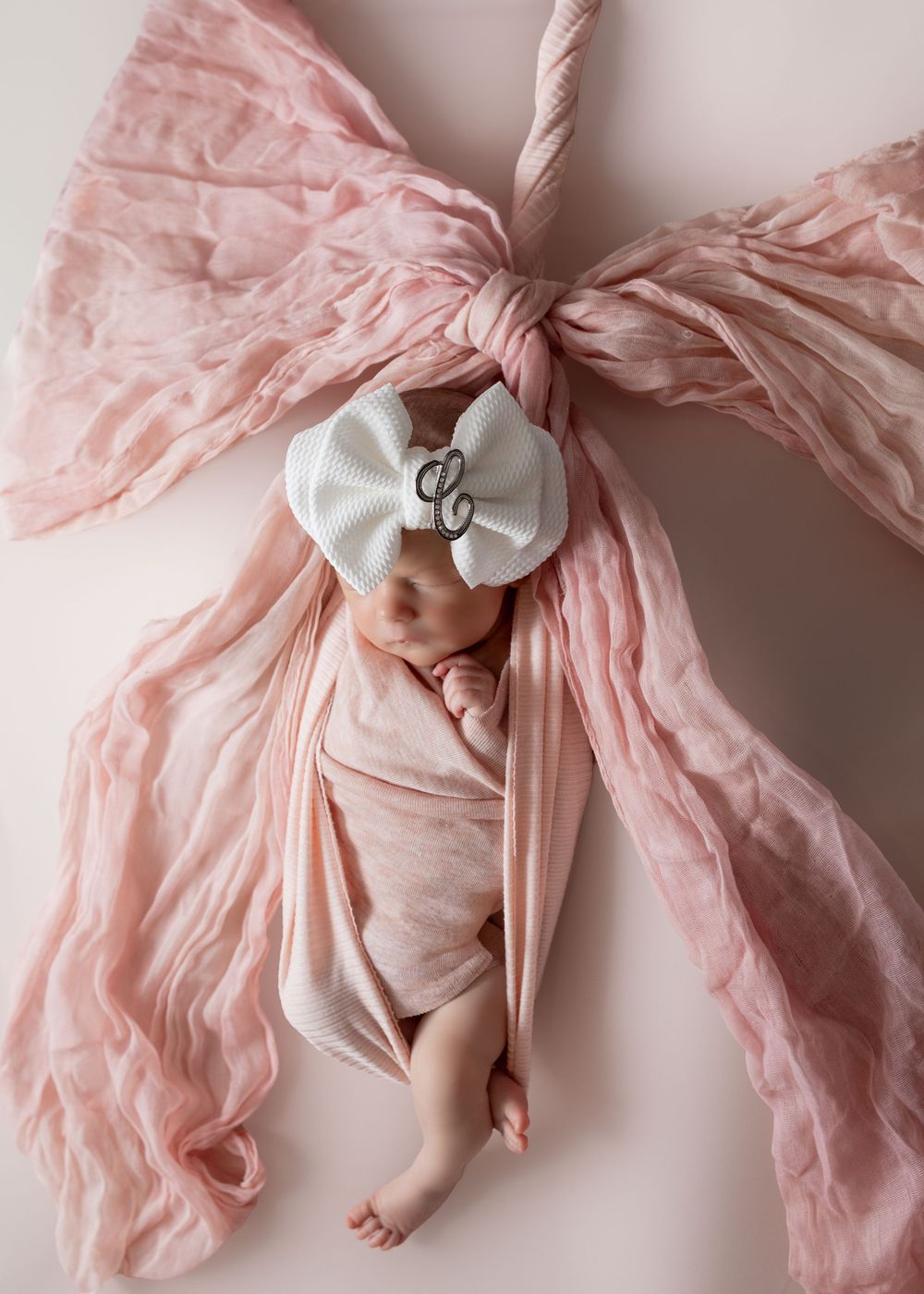 Newborn baby girl swaddled in pink wrap with white bow headband, posed on pink bow backdrop.