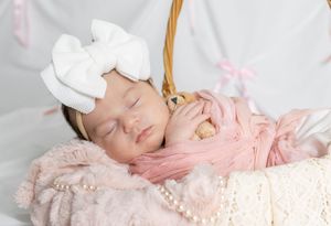 Peaceful newborn baby girl sleeping in basket wrapped in pink with white bow headband and pearl necklace.