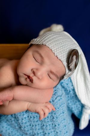 Sleeping newborn baby wearing a white knit hat with button detail, wrapped in a blue crochet blanket.