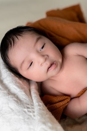 Peaceful newborn baby with dark hair resting on white lace and rust orange knit blankets.