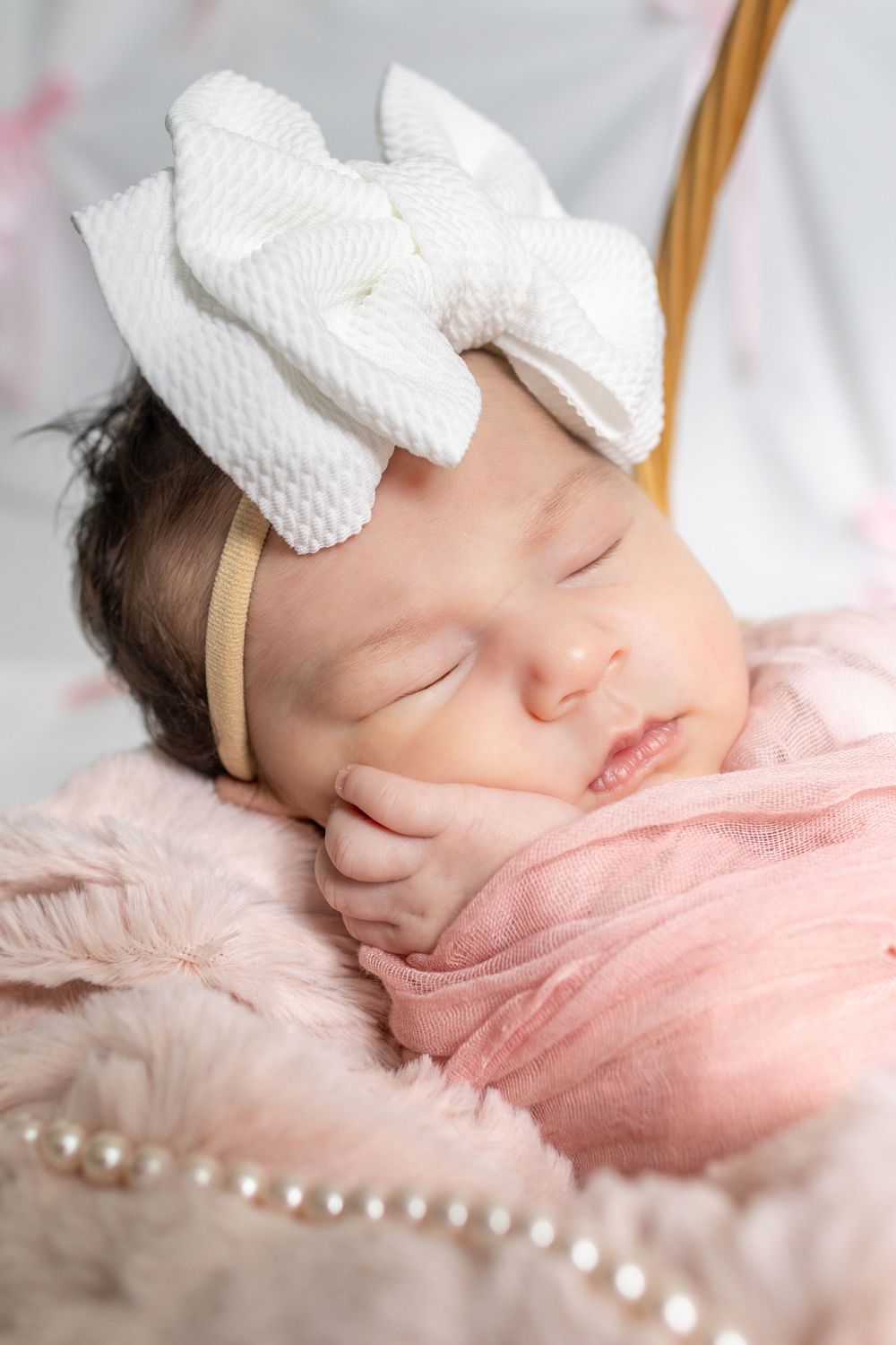 Sleeping newborn baby girl wearing a white bow headband, wrapped in a pink blanket with pearl accents.