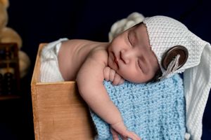 Sleeping newborn baby in white knit hat wrapped in blue blanket inside a wooden box.