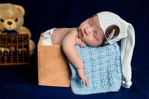 Newborn baby sleeping peacefully in a wooden box with a blue knit blanket and white hat, teddy bear in background.