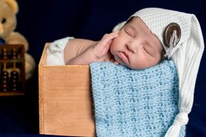 Newborn baby sleeping peacefully in a wooden box, wrapped in a blue knit blanket, wearing a white nightcap.