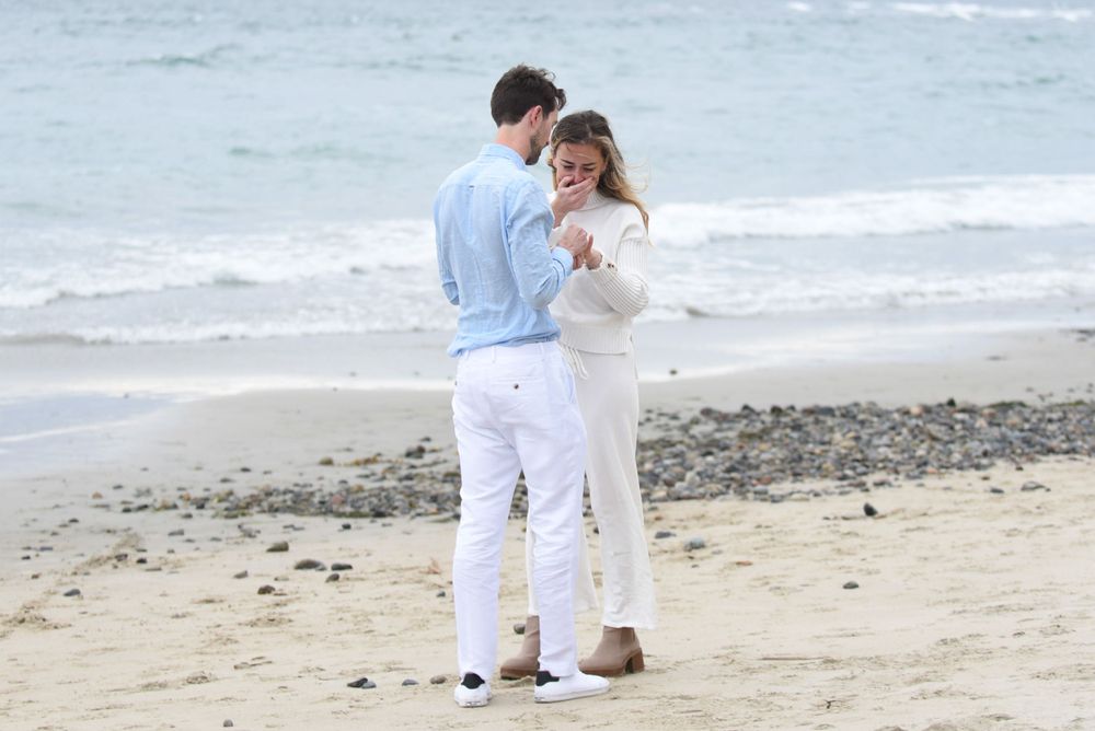 A couple in white and blue outfits embraces on a rocky beach with waves in the background.